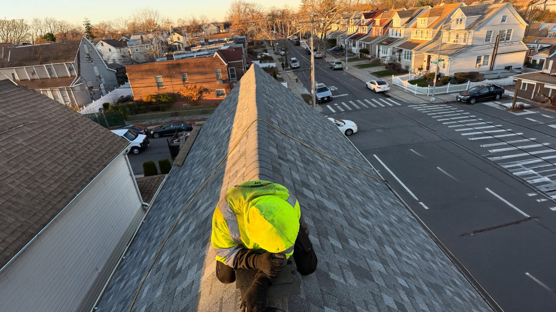 Roofer in a high-visibility yellow vest installing ridge cap shingles at the peak of a steep residential roof at sunset, with a dense urban street intersection and neighborhood row houses visible far below in Long Island.