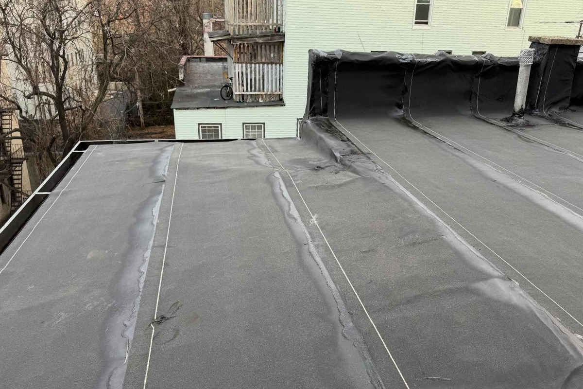 Flat urban rooftop with a deteriorating gray built-up roof showing seam cracks and ponding water stains, black EPDM membrane rolled back along the parapet walls, with neighboring green and yellow houses visible beyond.