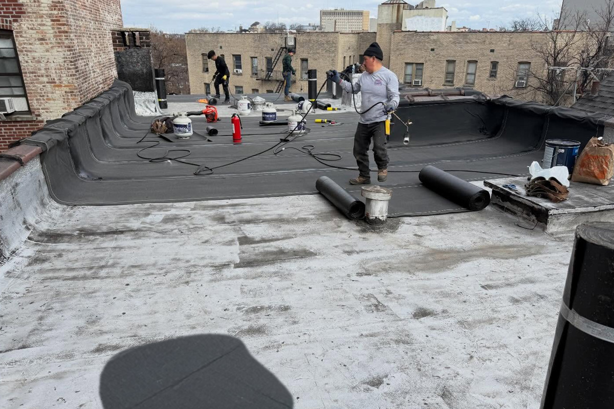 Three-person roofing crew installing EPDM rubber membrane on a urban flat roof, with rolled materials, adhesive buckets, and tools scattered across the surface, surrounded by brick apartment buildings.