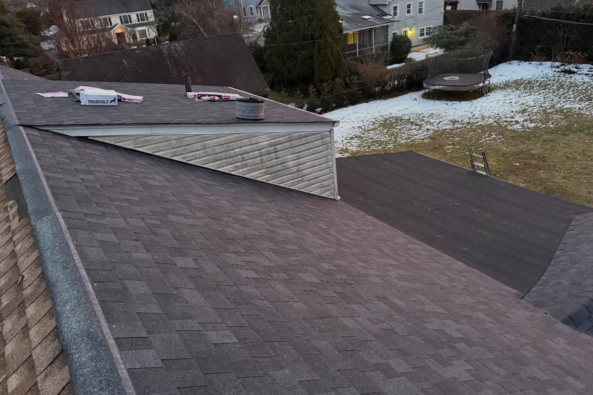 Aerial view of a multi-section residential roofing showing dark asphalt shingles, a flat rubber membrane section, a dormer with weathered siding, roofing underlayment materials on deck, and a snow-dusted suburban backyard below in Long Island.