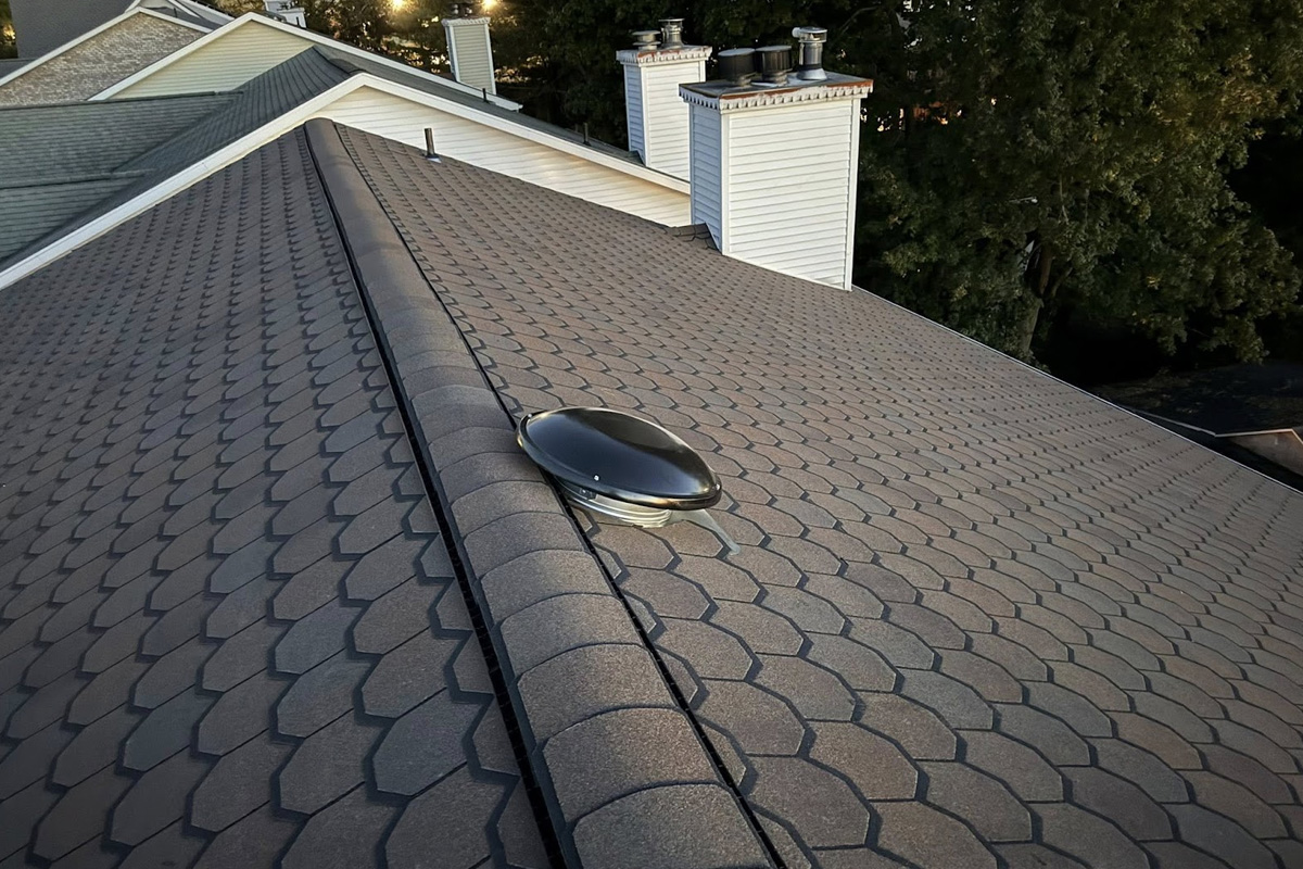 Shot displays brown scalloped shingles with a black solar attic fan. The ridge cap runs parallel to white chimneys and green trees, creating a clean, modern residential roofing profile in Long Island.