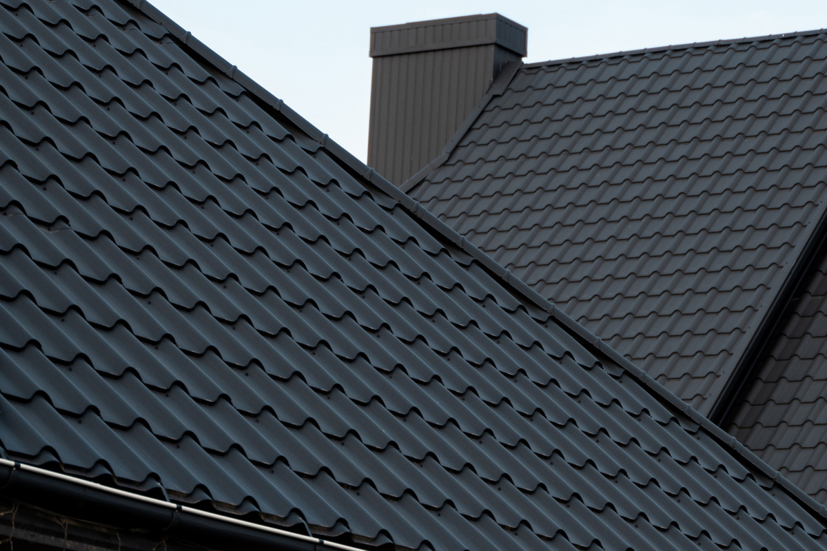 Close-up of roofing intersecting dark charcoal metal tile rooflines with a corrugated wave profile, a matching metal chimney cap, and a metal gutter visible along the eave under an overcast sky in Long Island.