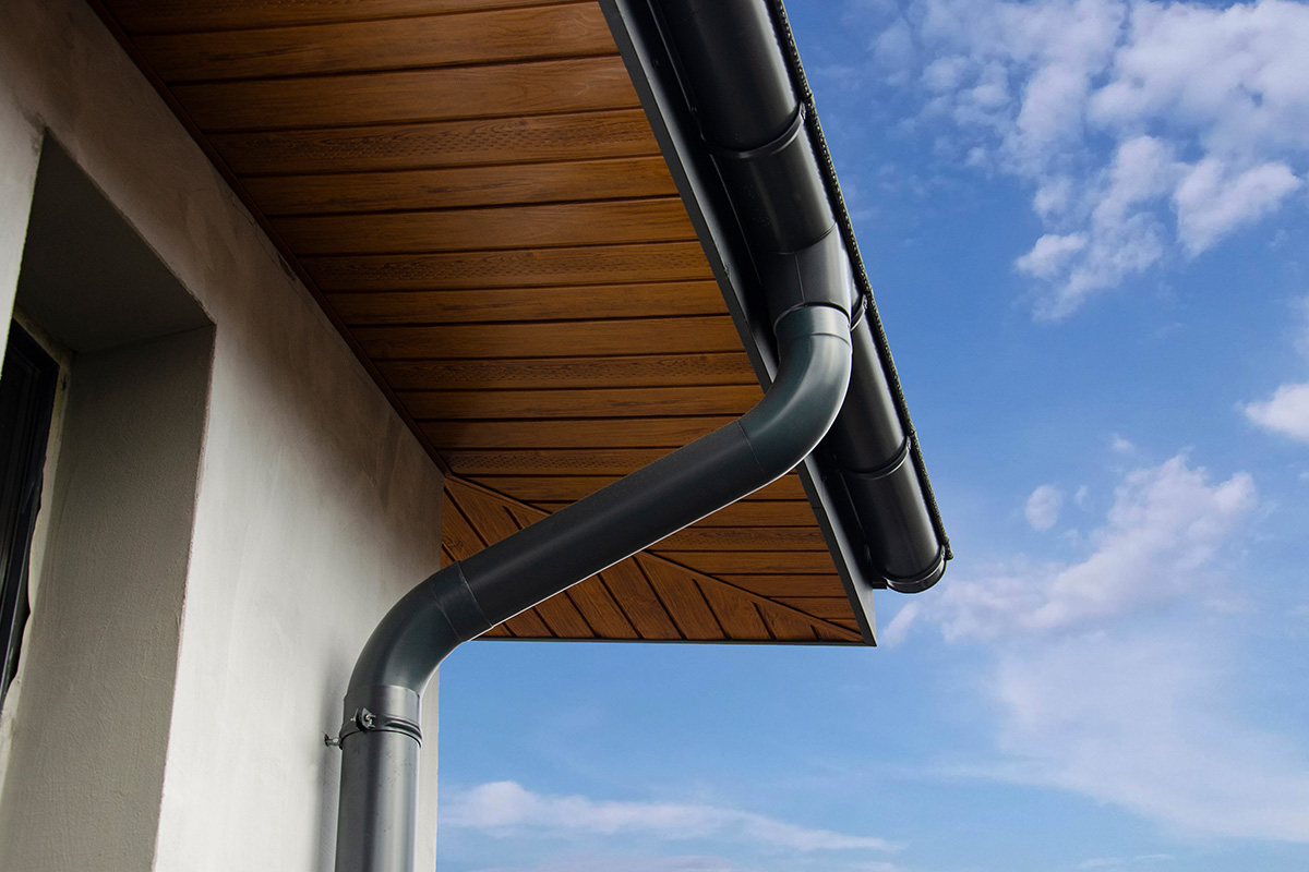 Close-up installation of a dark gray metal gutter and round downspout with elbow fitting mounted on a home with a warm wood-paneled soffit, white stucco wall, and blue sky with clouds in the background.