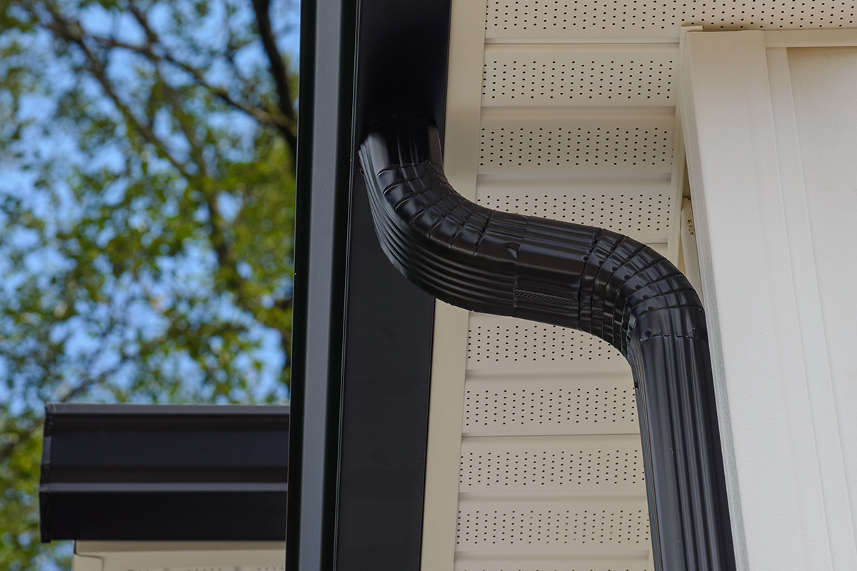 Close-up installation of a black aluminum downspout with offset elbows mounted against beige perforated vinyl soffit and white fascia, with a black gutter and green tree foliage visible in the background.