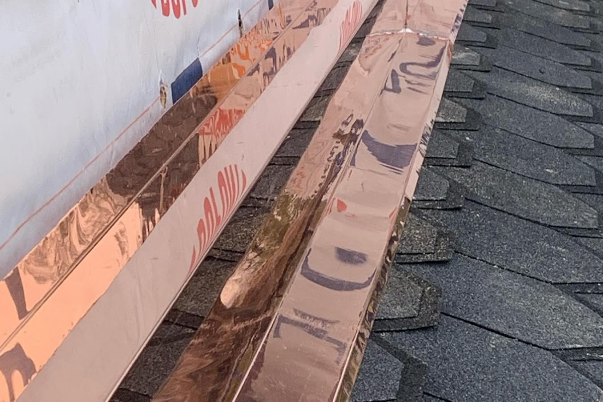 Image features a close-up of a dark grey shingled roof during a construction or repair project. Long strips of polished copper flashing are laid out across the shingles, reflecting the surrounding environment on their metallic surface in Long Island.