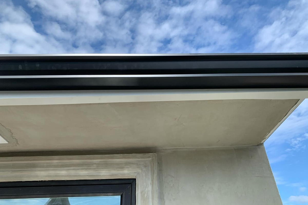 Image shows a close-up of a residential roofline, featuring a modern black gutter system installed against a white soffit. Below the roof edge, a portion of a black-framed window is visible, surrounded by a light-colored, textured exterior wall in Long Isalnd.