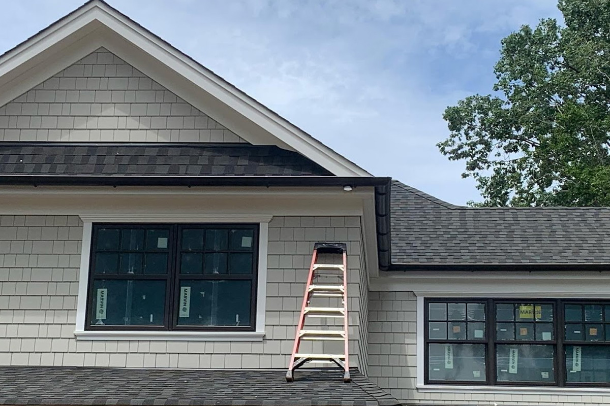 A light grey shingled house features dark-framed windows with white trim and a black gutter system. An extension ladder leans against the exterior during this ongoing installation in Long Island.