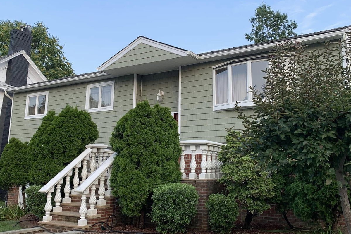 A residential home features horizontal green shingle siding across the front facade. This green siding is accented by white window trim and a white-pillared staircase in Long Island.