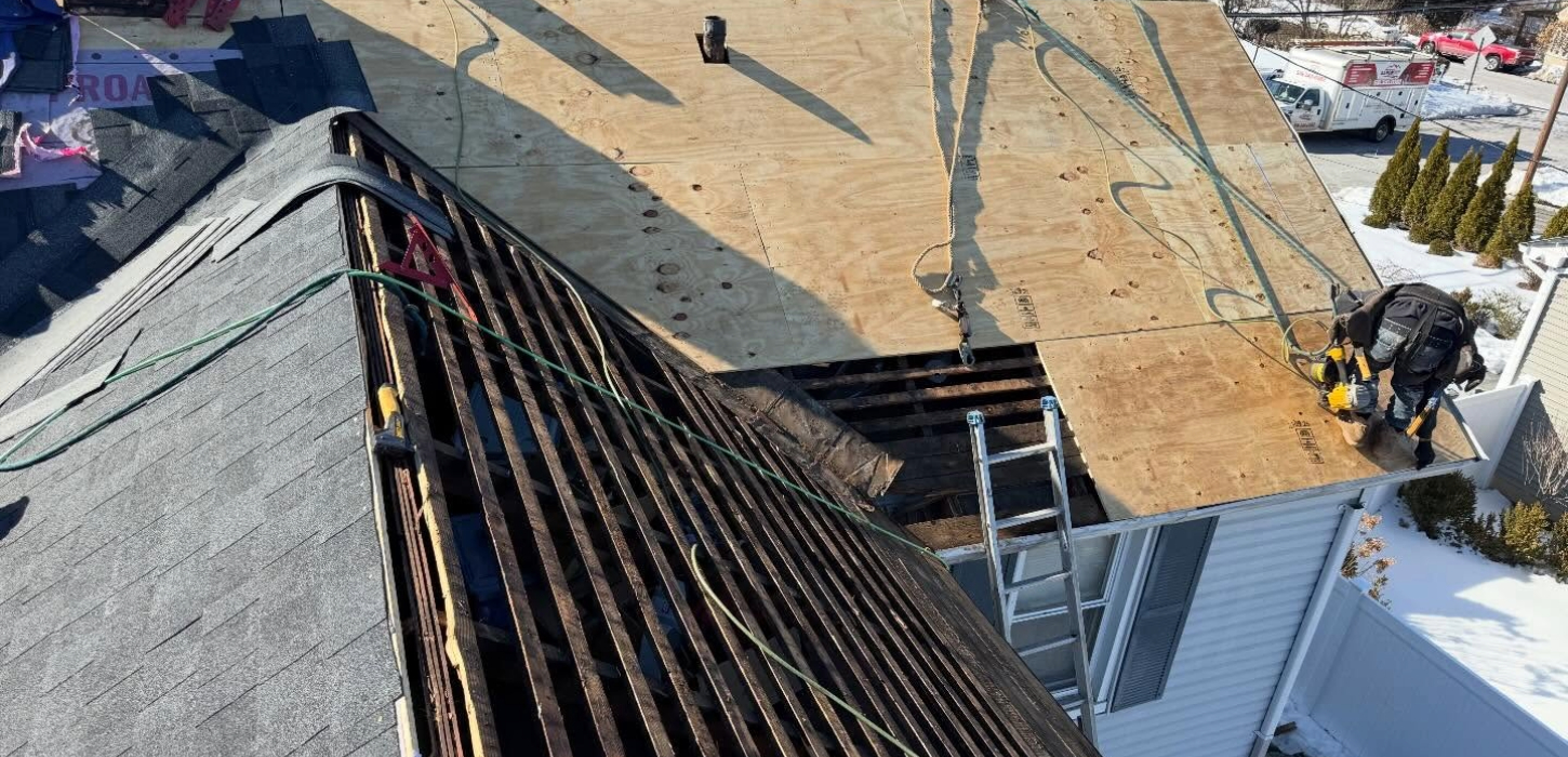 Workers during roof replacement install new plywood decking over exposed wooden slats on a residential roof. A technician uses power tools while safety ropes and a ladder facilitate the shingle replacement in Long Island.