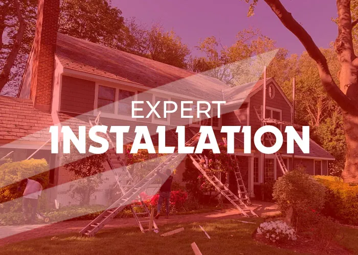 Workers perform a residential roofing installation using ladders and scaffolding. The gray-sided house features a brick chimney, while green grass and flowering bushes landscape the property in Long Island.