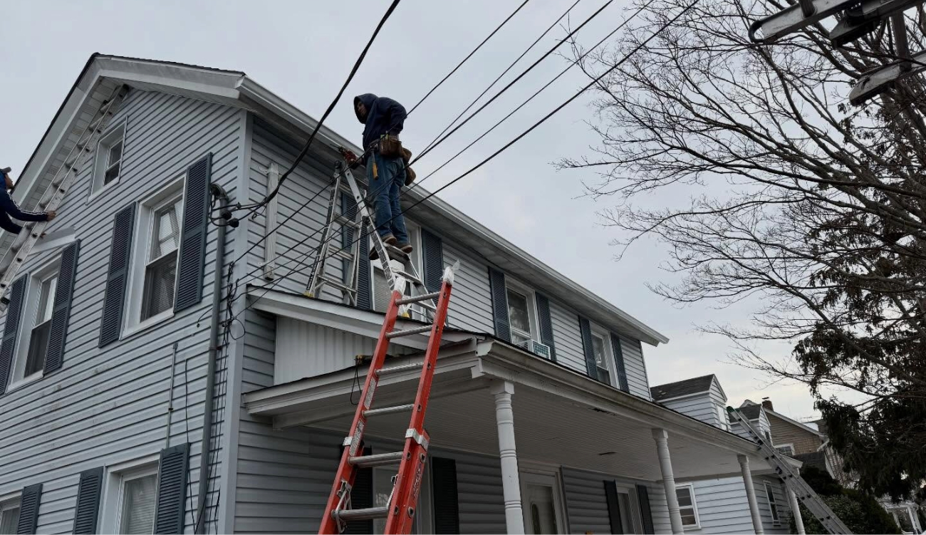 A roofer on a ladder working on the edge of a two-story house performing roof flashing installation.