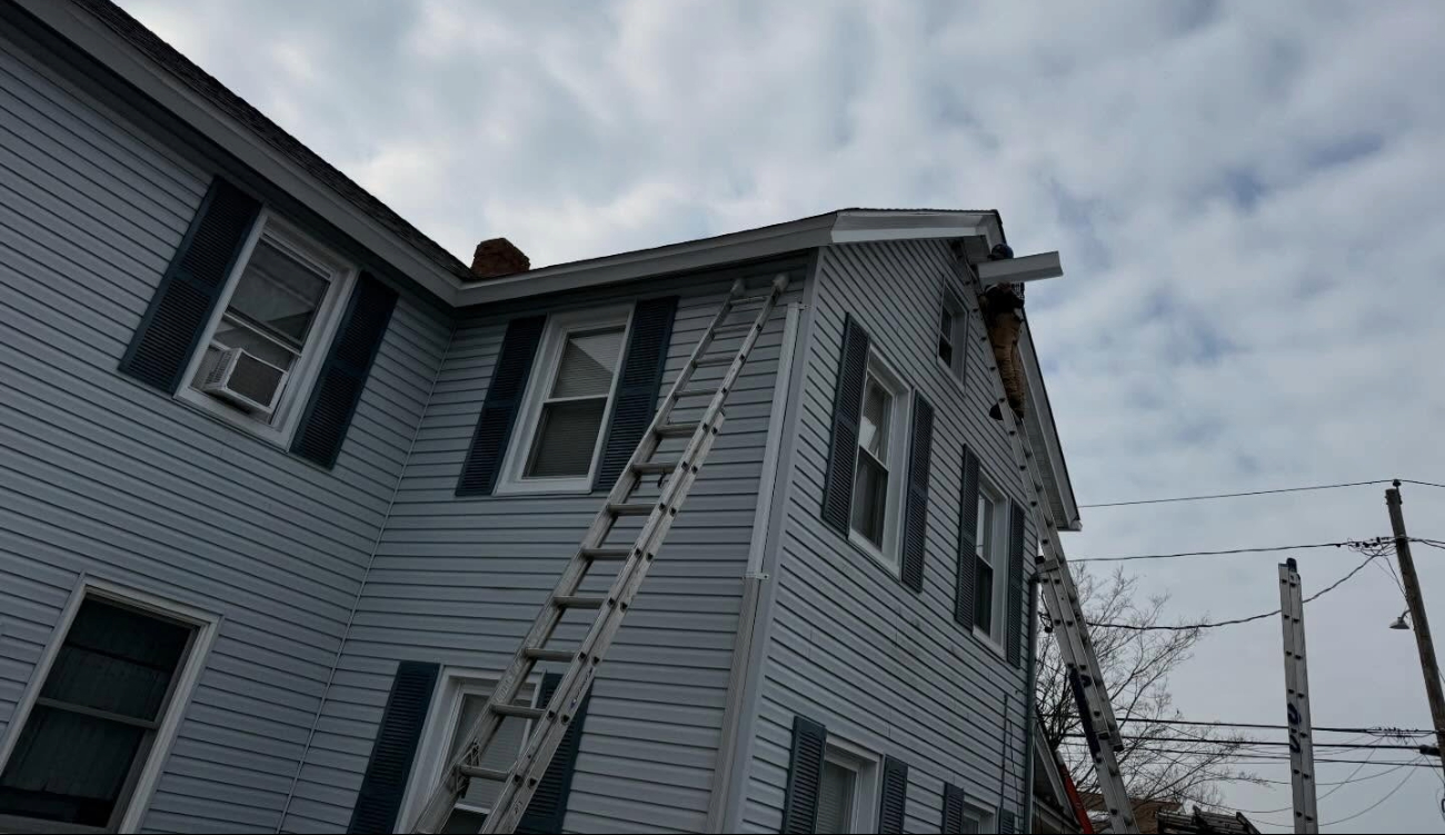 A roofer on an extension ladder working on the gable end during a roof flashing installation.