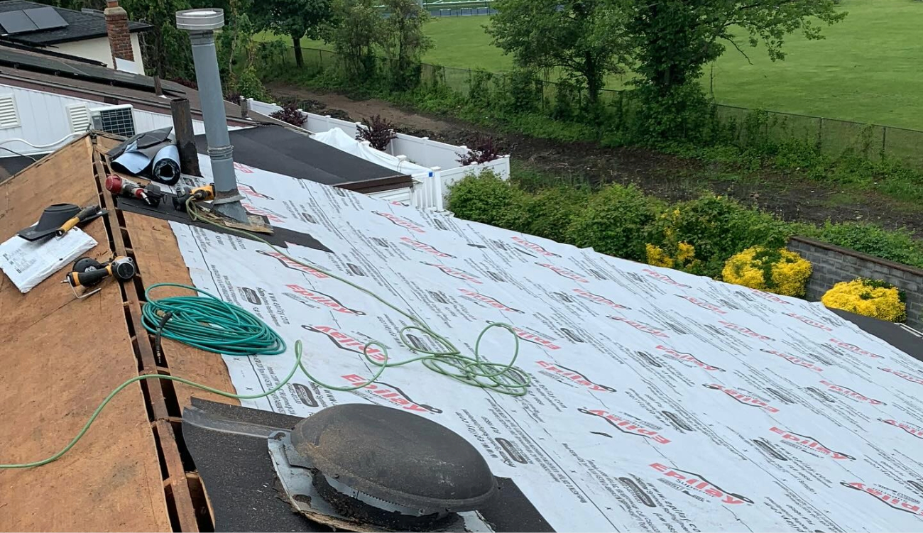 A close-up of a residential roof deck covered with underlayment, featuring a dome vent prepared for a roof ventilation installation.