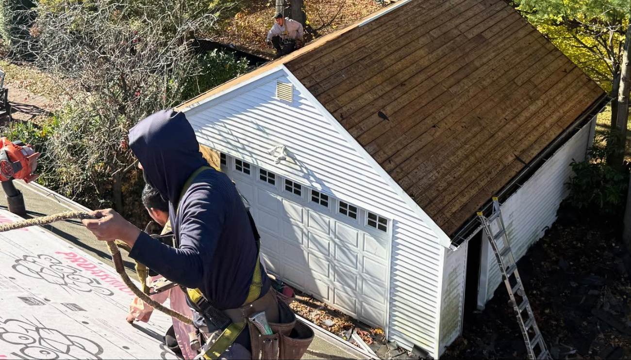 Roofers in safety harnesses preparing a home's roof deck and nearby garage for new materials and a proper roof ventilation installation.