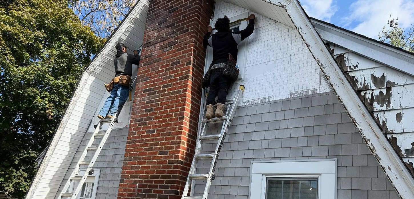 Workers on ladders repairing a house exterior during a storm damage roof replacement Nassau County project.